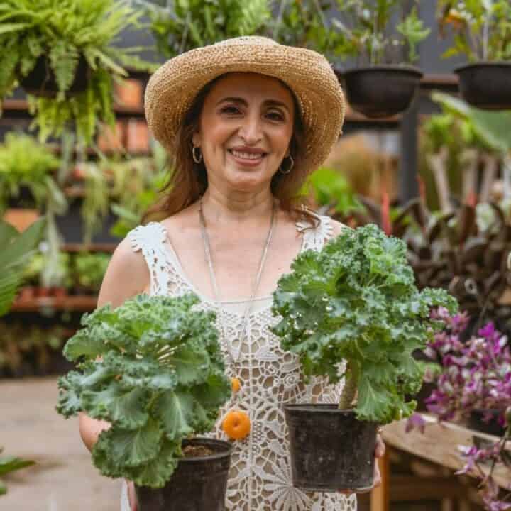A woman in a straw hat holds two potted leafy plants, standing in a garden center surrounded by various plants and flowers.
