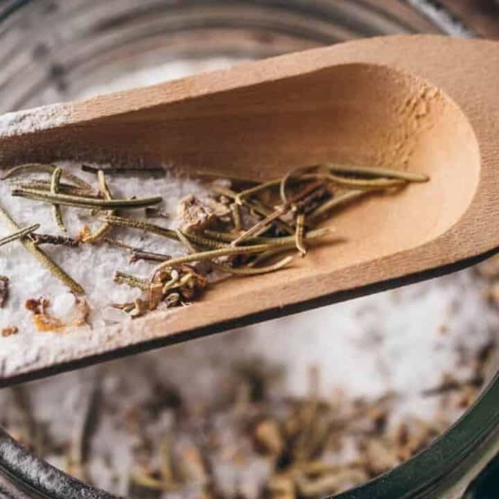 Wooden scoop with coarse salt and dried rosemary above a glass jar.