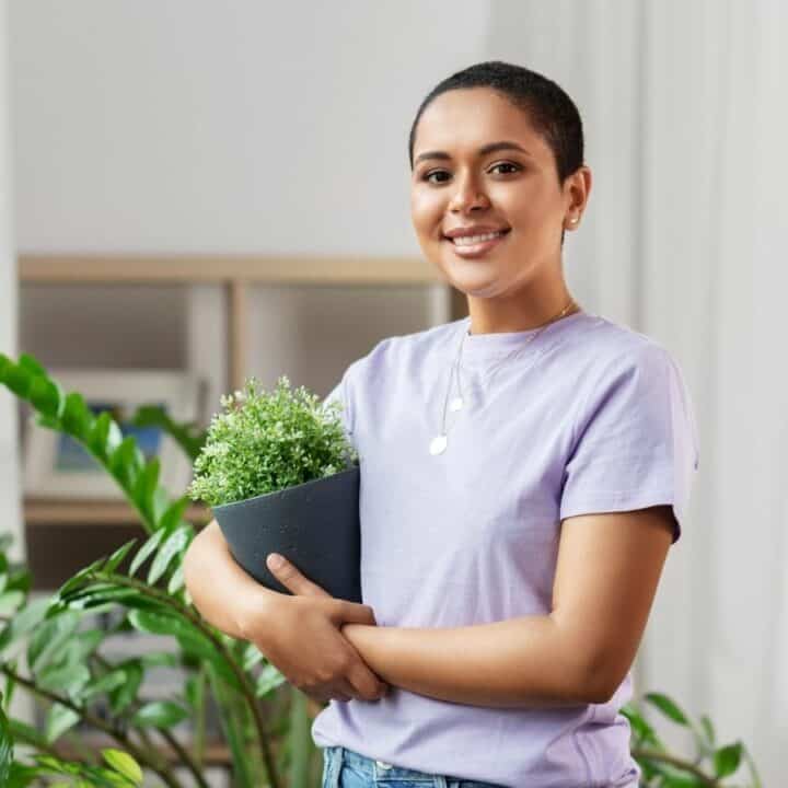 Person in a lavender shirt holding a potted plant and smiling, surrounded by other plants indoors.