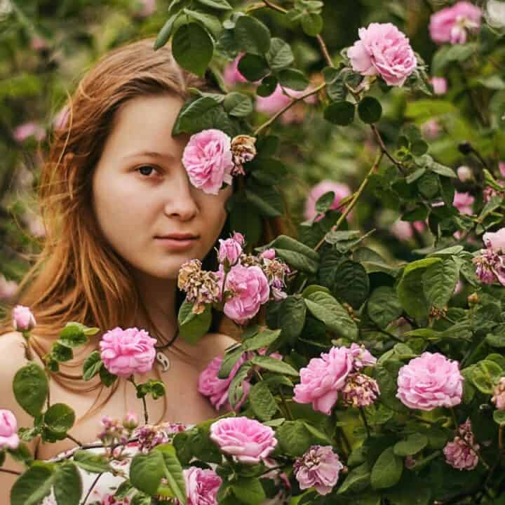 A woman stands partially obscured by dense pink rose bushes, looking directly at the camera.
