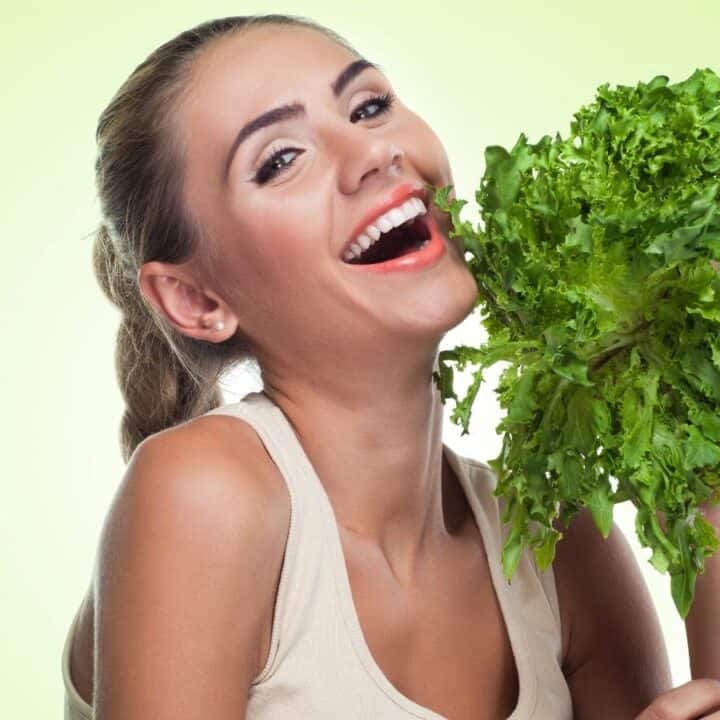 A smiling woman in a sleeveless top holds a bunch of leafy green lettuce in front of a light green background.
