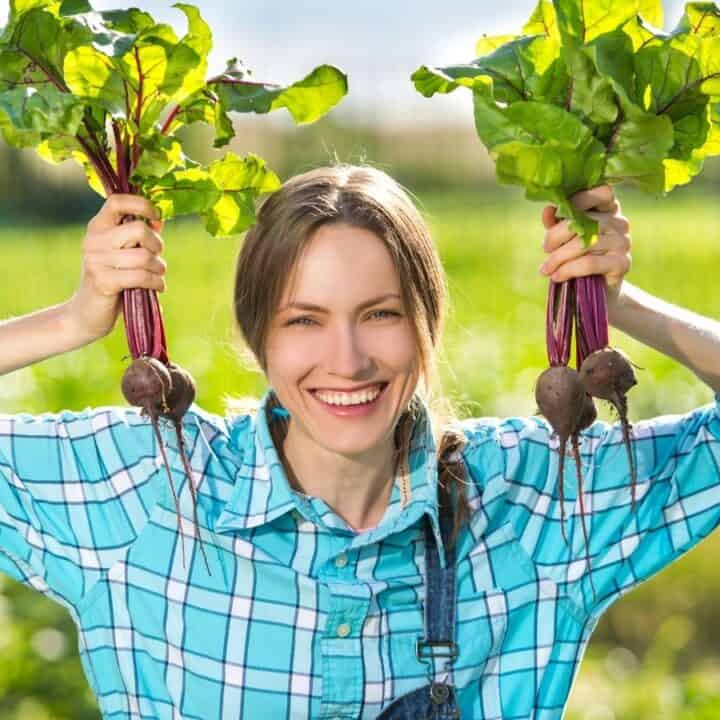 A woman in a plaid shirt smiles and holds up two bunches of freshly harvested beets in a garden.