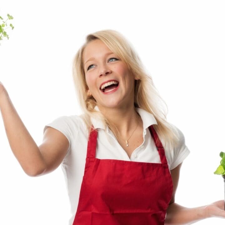 Smiling woman in a red apron holds two potted green plants, one in each hand, against a white background.