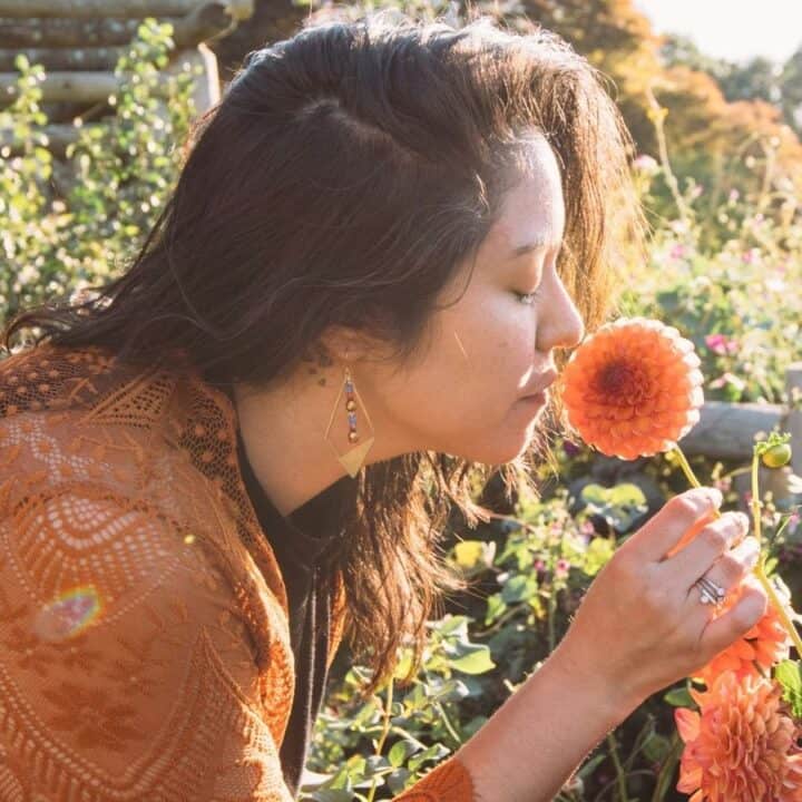 A person wearing an orange cardigan is smelling a large, blooming flower in a garden with greenery in the background.