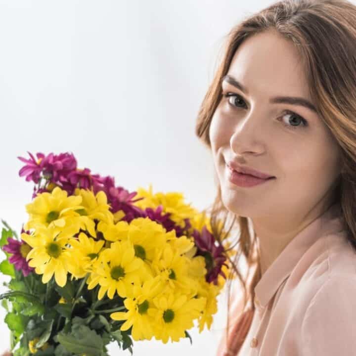 A woman with long brown hair holds a bouquet of yellow and purple flowers, looking at the camera and smiling slightly.