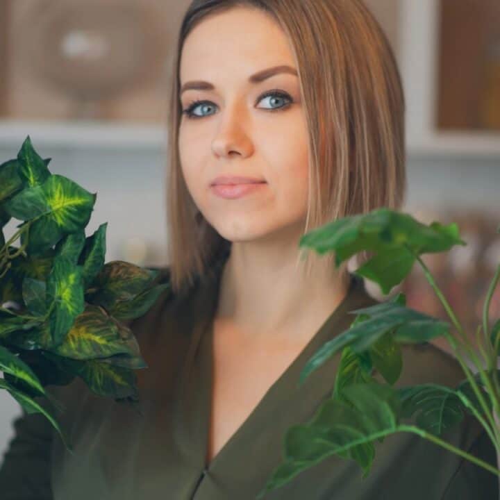 A woman holding a potted plant in each hand, standing indoors with blurred shelves in the background.