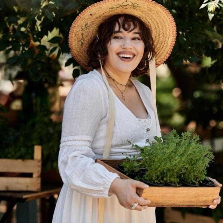A woman in a straw hat and white dress holds a wooden box of green plants outdoors, smiling at the camera.
