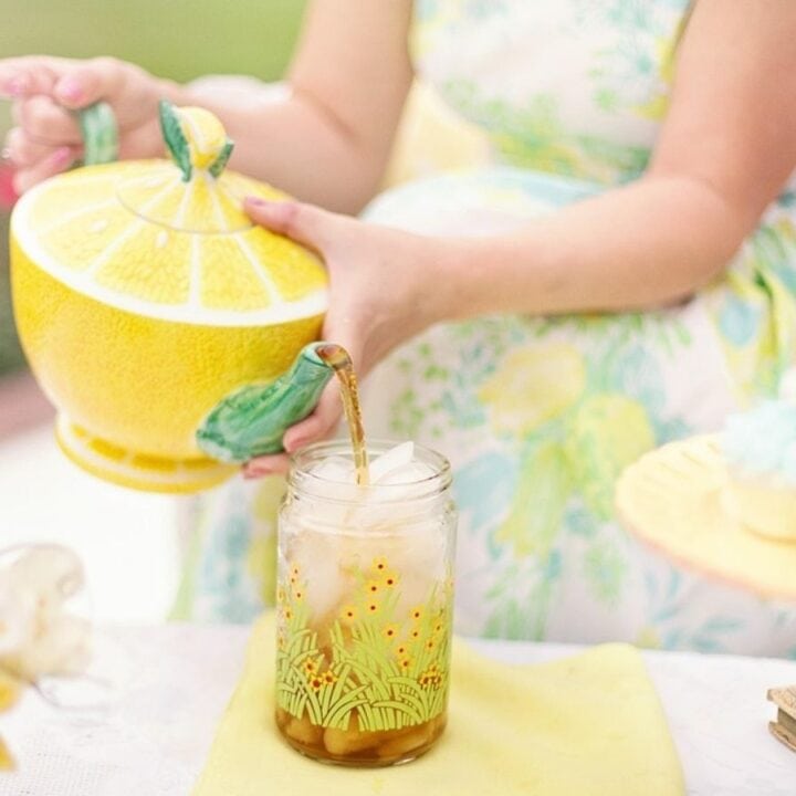Person pouring iced tea from a lemon-shaped teapot into a jar. A plate with blue cupcakes is on the table, surrounded by flowers.