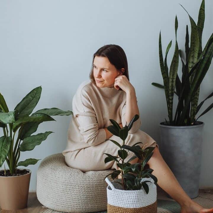 A woman in a beige dress sits on a pouf indoors, surrounded by three potted plants against a light-colored wall.