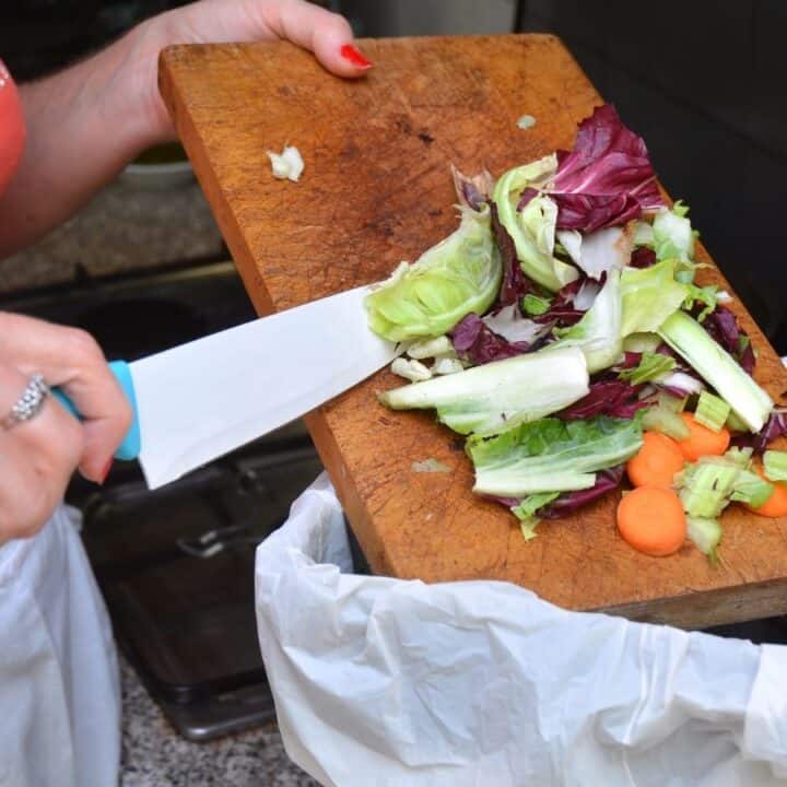A person uses a knife to scrape vegetable scraps from a cutting board into a trash bin lined with a plastic bag.