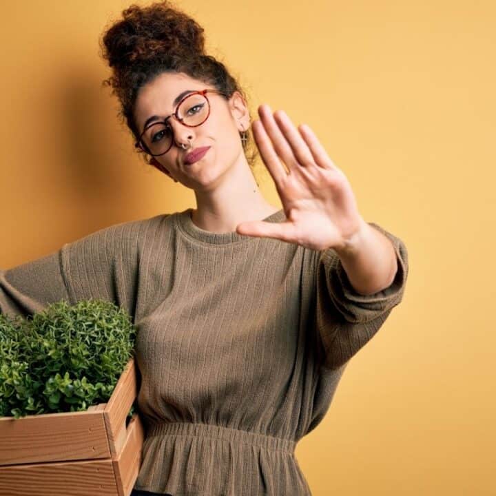 A woman holding a wooden box of green plants raises her hand palm-forward in a stop gesture, standing against a plain yellow background.