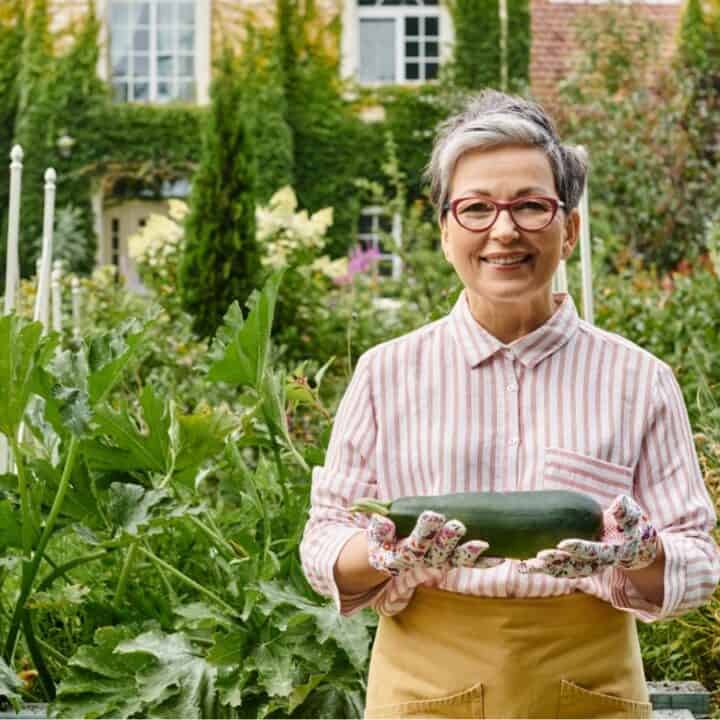 A woman stands in a lush garden holding a large zucchini, with a house covered in ivy visible in the background.