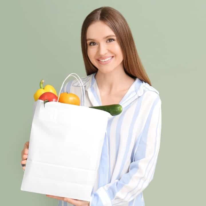 A woman with long brown hair in a striped shirt holds a white bag filled with various vegetables against a light green background.