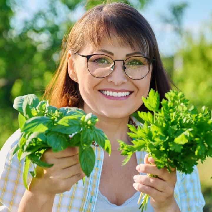 A woman wearing glasses stands outdoors, smiling and holding bunches of fresh green herbs in each hand.