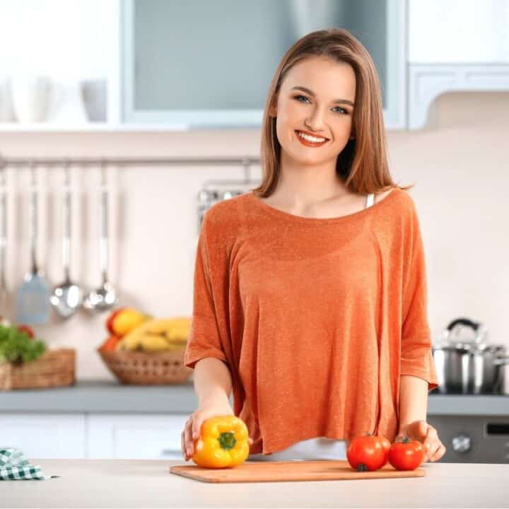 Woman smiling in a kitchen while holding a yellow bell pepper and standing next to tomatoes on a cutting board. Cooking utensils and vegetables are visible in the background.
