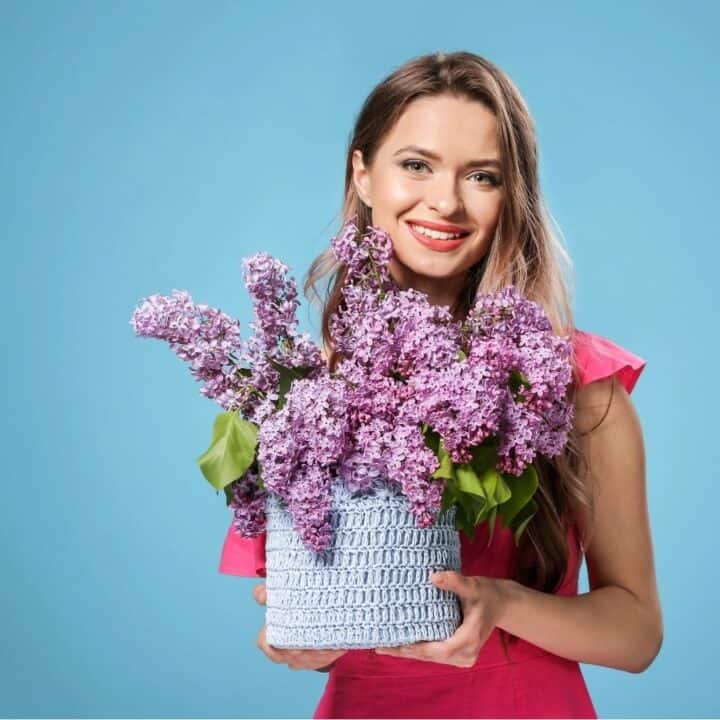 A woman in a pink dress holds a basket of purple lilacs against a blue background, smiling at the camera.