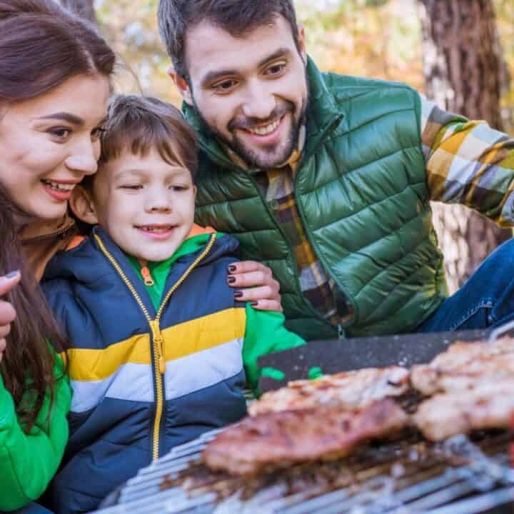 Family of three, dressed in autumn jackets, smiling and grilling food on a red barbecue in a wooded area.