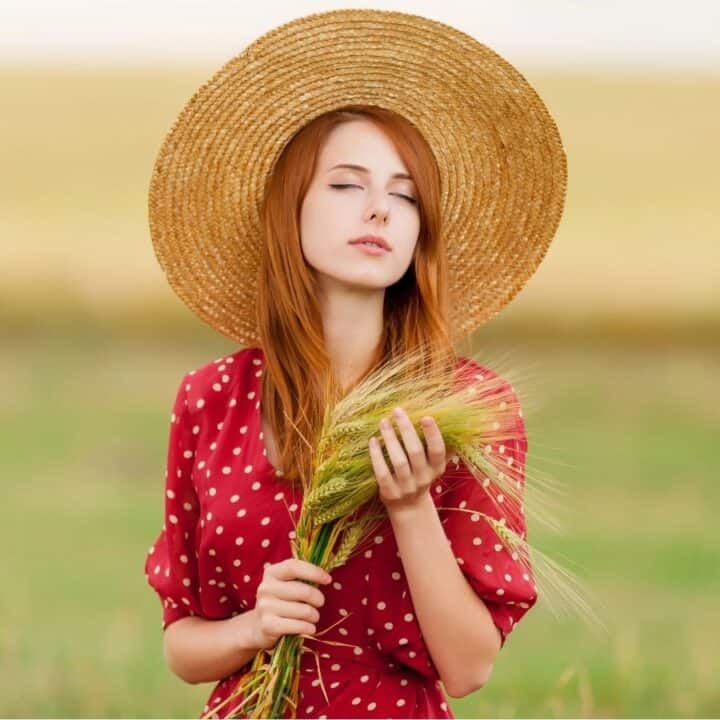 A woman in a red polka dot dress and wide straw hat holds a bundle of wheat while standing in a green field.