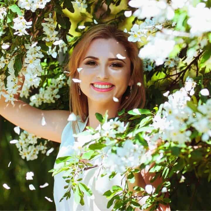 A woman stands outdoors among blooming white flowers, smiling at the camera with one arm raised, surrounded by green foliage and falling petals.
