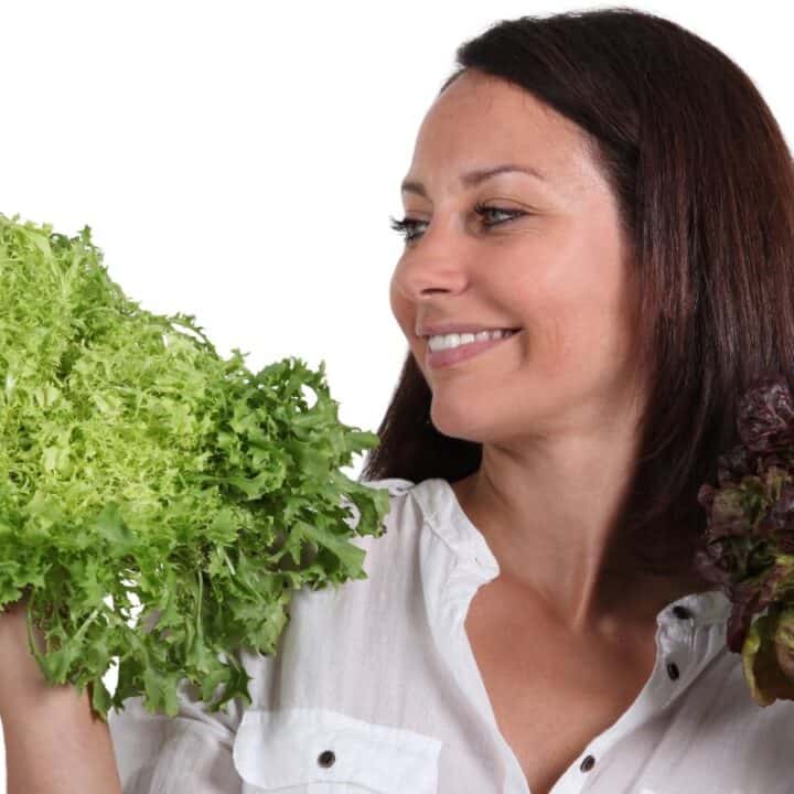 A woman in a white shirt holds up a bunch of green leafy lettuce in one hand and a bunch of purple-red lettuce in the other, smiling and looking at the green lettuce.