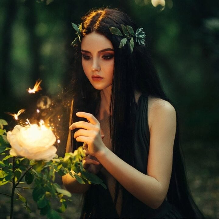 A woman with long dark hair and a leafy headpiece touches a glowing white flower in a lush, green forest setting.