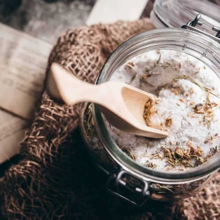 A glass jar filled with coarse bath salts and dried herbs, with a wooden scoop, rests on a burlap surface beside open, vintage-style books.