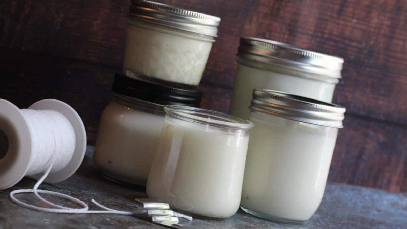 Five jars of white wax sit on a surface, with a spool of wick and metal wick clips nearby.