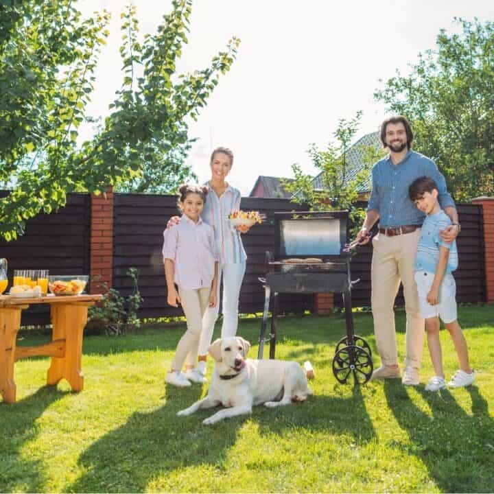 A family of four and a dog stand beside a grill in a sunny backyard with food and drinks on a nearby table.