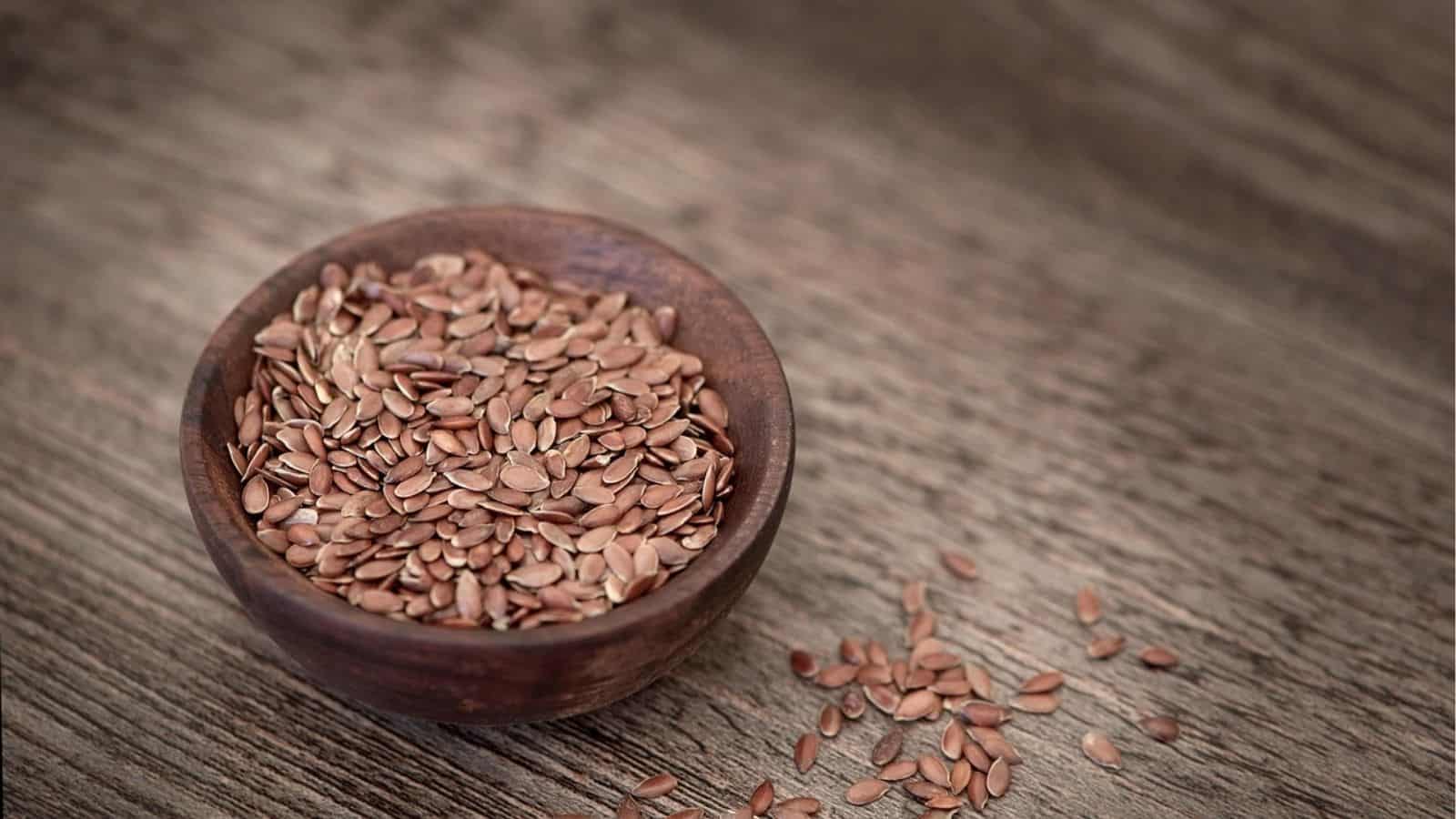 A wooden bowl filled with flaxseeds on a textured wooden surface. Some seeds are scattered outside the bowl.