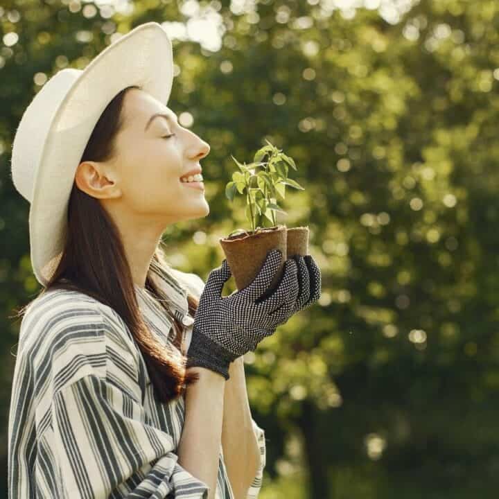 Woman in a hat smiles and holds a small potted plant outdoors, surrounded by greenery.