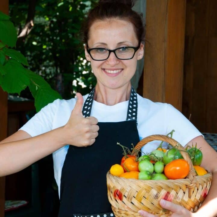 A woman wearing glasses and an apron holds a basket of assorted tomatoes and gives a thumbs up while standing outdoors.