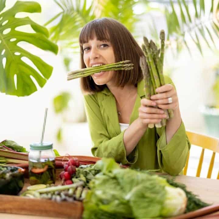 Woman sitting at a table with assorted vegetables, holding and biting a bunch of asparagus, surrounded by leafy greens and a drink jar.