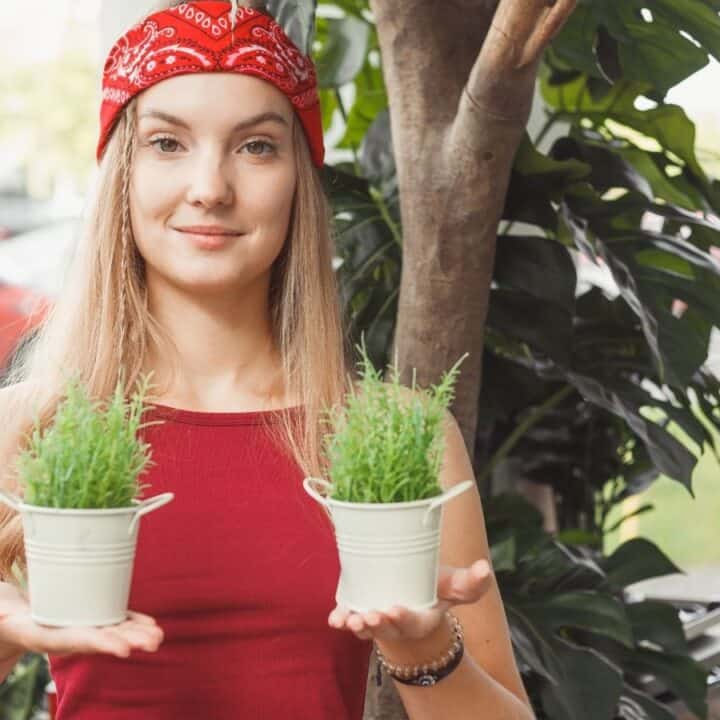 A woman wearing a red bandana and red top holds two small potted plants, standing outdoors among greenery.