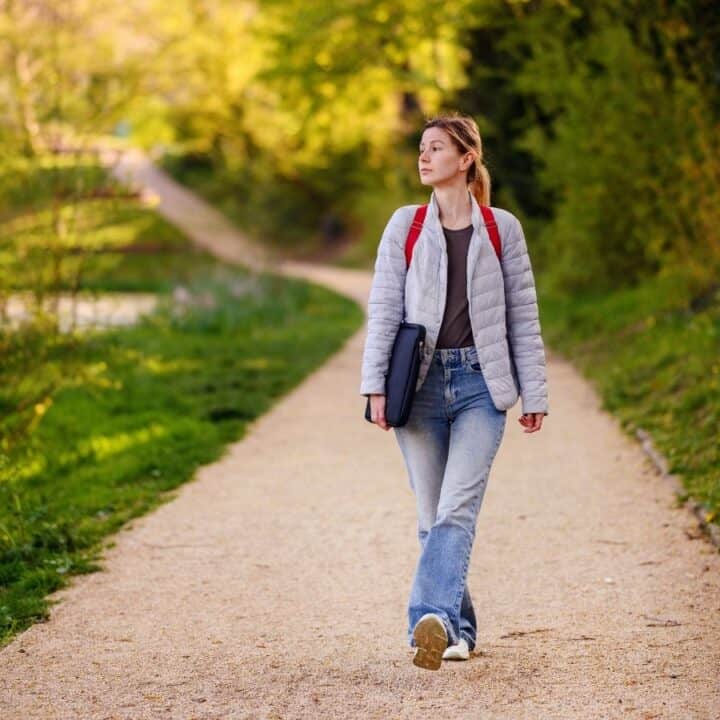 A person with a red backpack walks alone on a gravel path through a green, wooded area, holding a laptop or book under one arm.