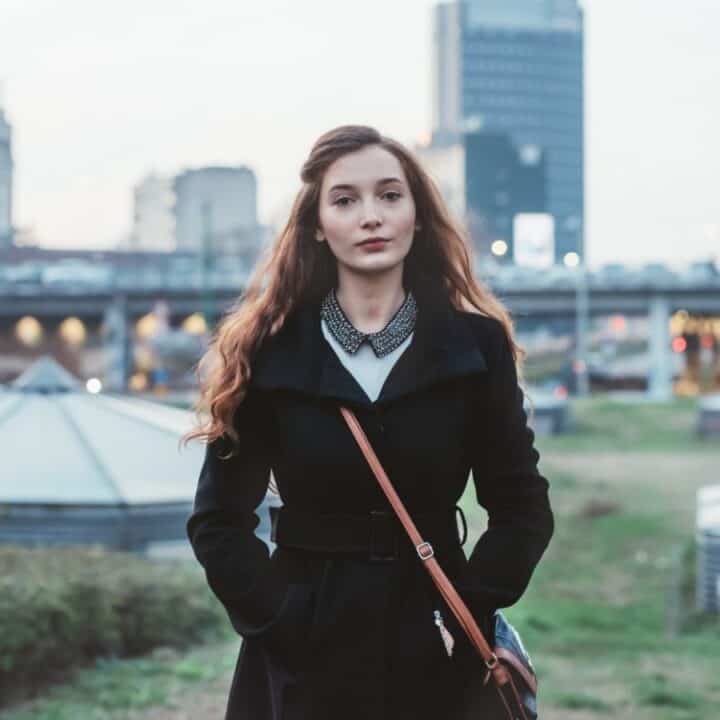 A woman with long red hair wearing a black coat stands outdoors with her hands in her pockets; a cityscape and buildings are visible in the background.