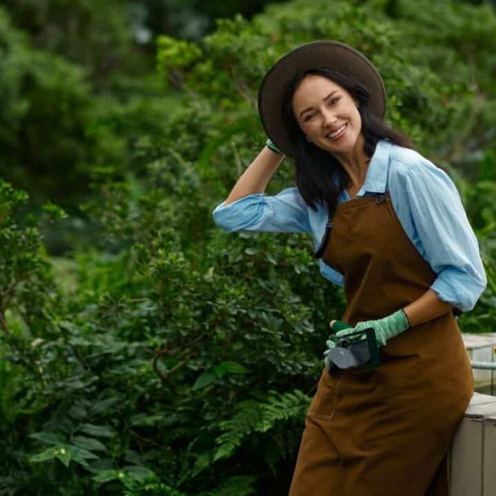 A woman wearing a brown apron, gloves, and hat stands in a garden, smiling with one hand behind her head. A yellow watering can is on the ledge next to her.