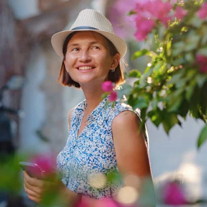 A woman wearing a white hat and blue floral dress smiles while standing outdoors near pink flowers, holding a smartphone.
