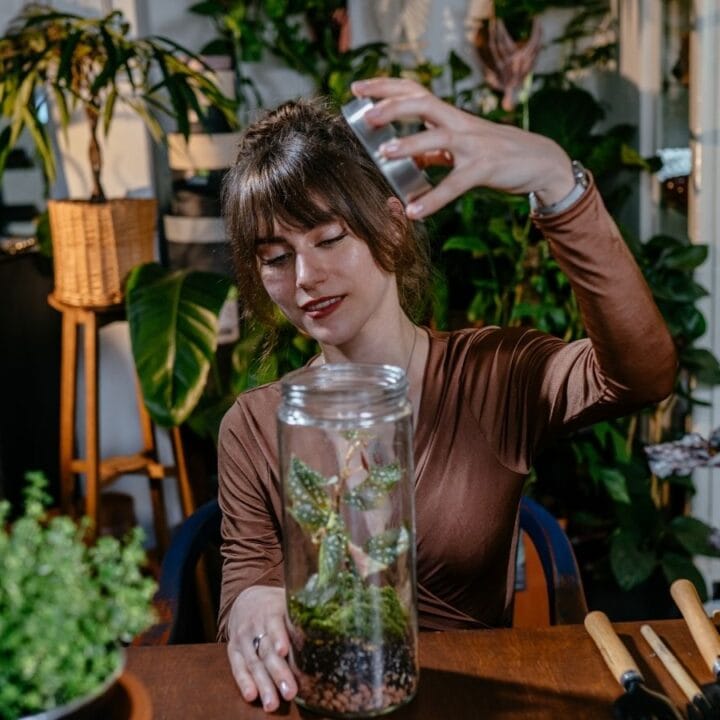 A woman sprays water into a glass jar containing a small plant while sitting at a wooden table surrounded by various houseplants.