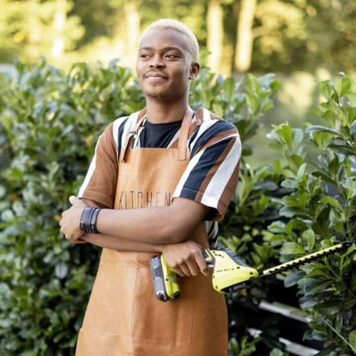 A person wearing a brown apron stands outdoors in front of green bushes, holding a hedge trimmer and smiling with arms crossed.