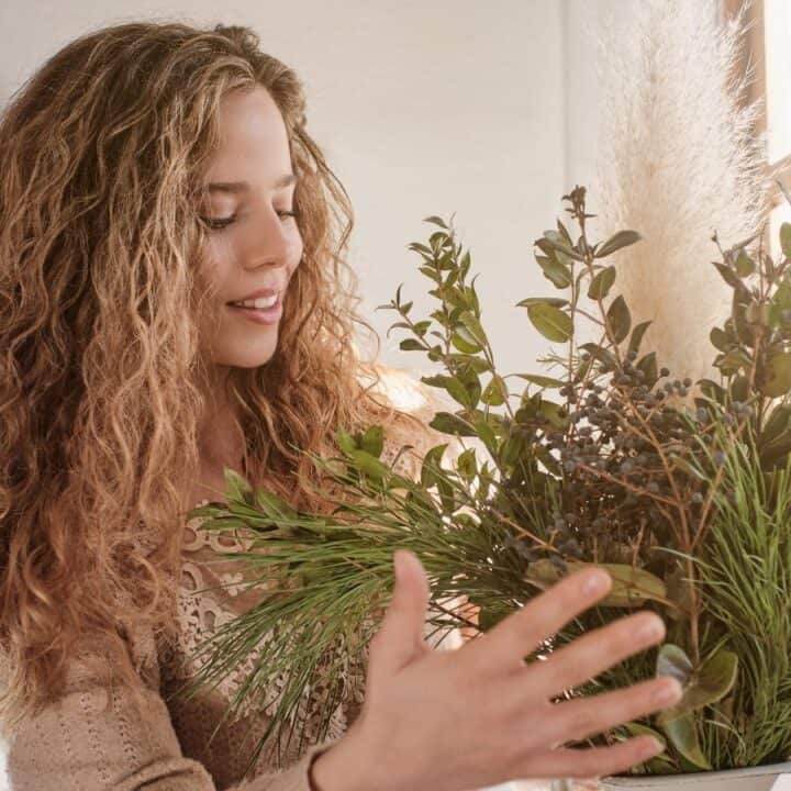 A woman with curly hair stands indoors by a window, holding and looking at a large arrangement of green foliage and branches.