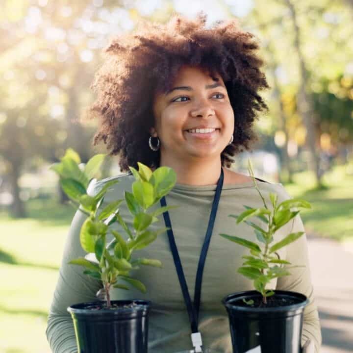 A person outdoors in a park carrying two small potted plants, smiling and wearing a lanyard.