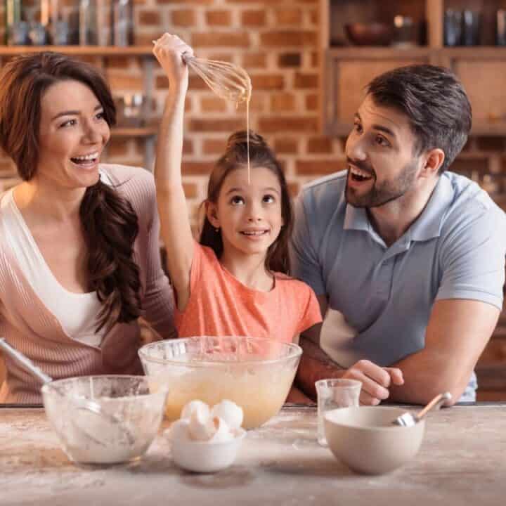 A child holds up a whisk in a kitchen while two adults smile beside her; baking ingredients and utensils are on the counter.
