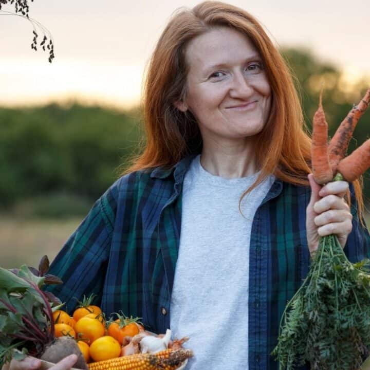 A woman standing outdoors holds freshly harvested vegetables, including carrots, tomatoes, and corn, and smiles at the camera.