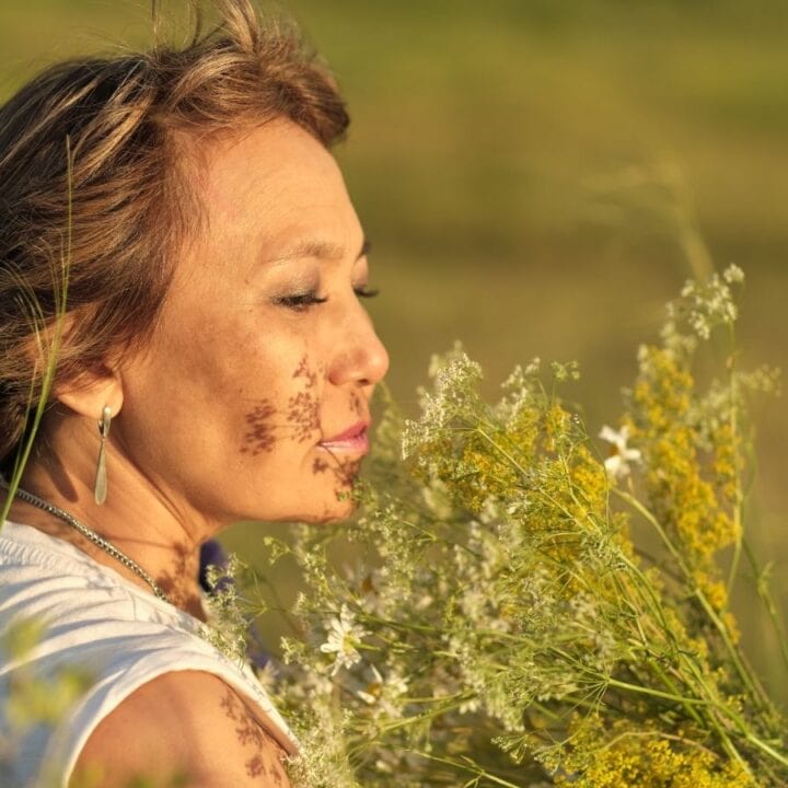 A person with short hair stands outdoors in a field, holding and smelling a bouquet of wildflowers, with sunlight illuminating their face.
