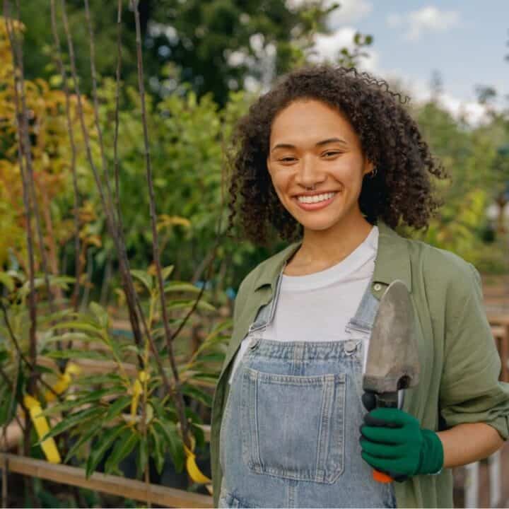A woman wearing overalls, gloves, and a green shirt stands in a garden holding a trowel, surrounded by plants and trees on a sunny day.