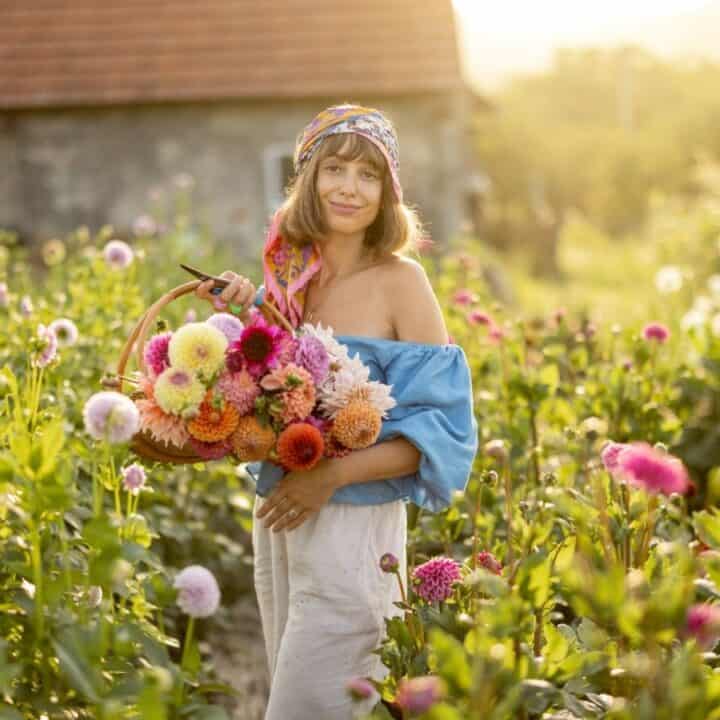 A woman standing in a flower field holds a basket of colorful flowers, wearing a blue off-shoulder top, white skirt, and a headscarf. A rustic building is in the background.