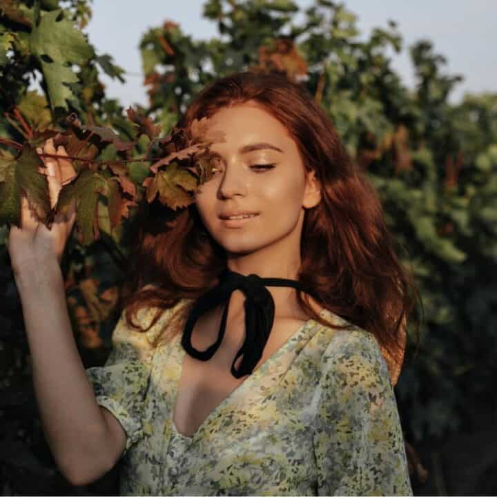 A woman with long red hair stands in a vineyard, gently touching grapevine leaves, wearing a floral dress with a black ribbon at the neck.
