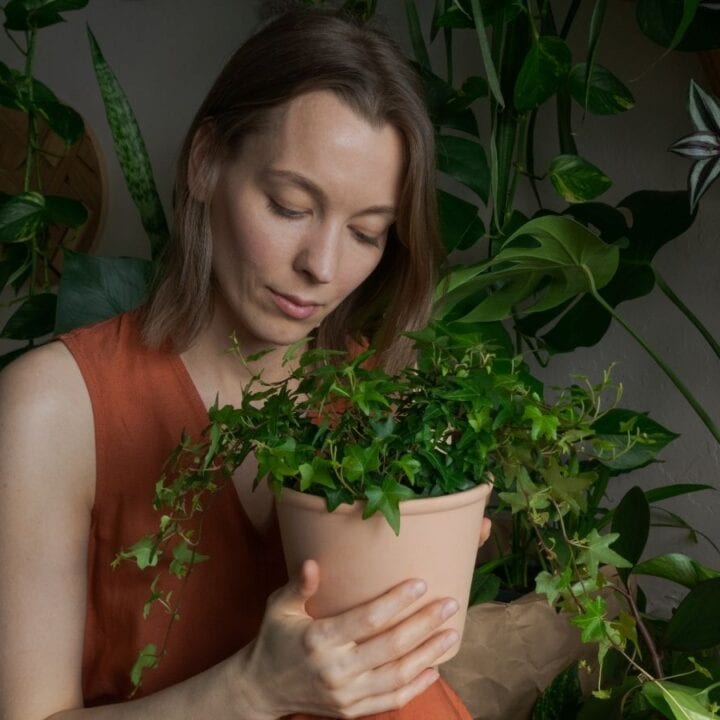 A woman in an orange sleeveless top holds and looks at a potted plant, surrounded by various green houseplants.
