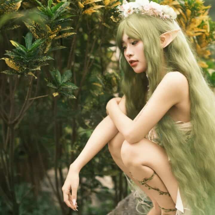 A person in a fantasy elf costume with long green hair, pointed ears, and a flower crown, crouches beside green plants outdoors.