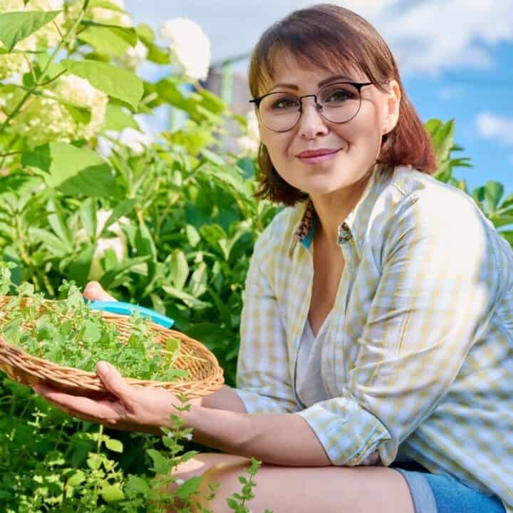 A woman wearing glasses holds a wicker basket with fresh herbs while kneeling in a lush garden on a sunny day.